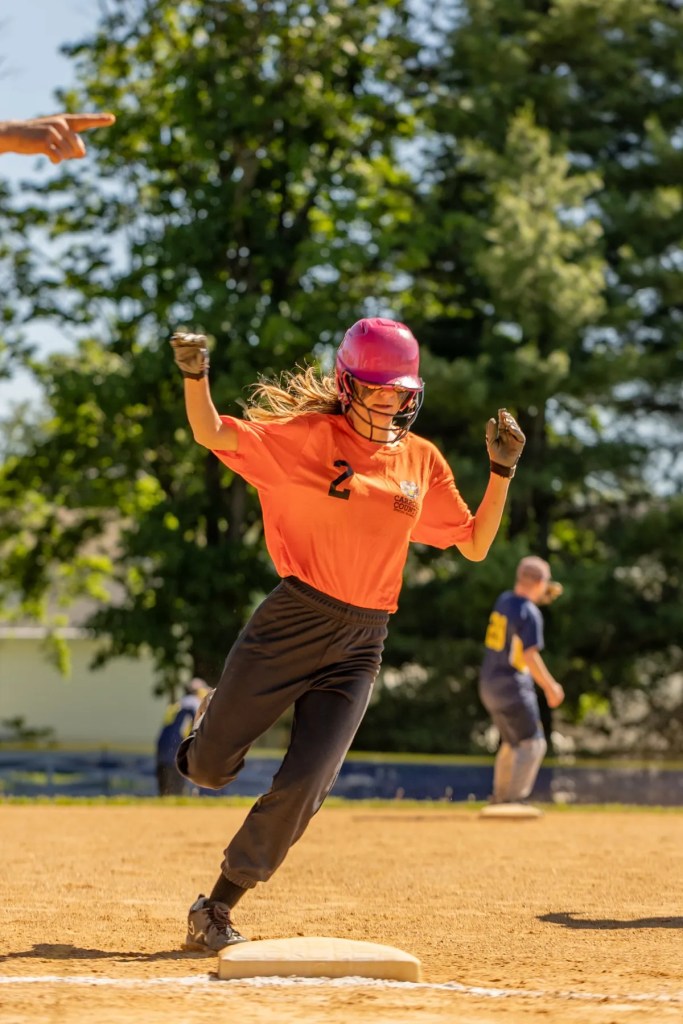female softball player in pink helmet and orange uniform running past a base